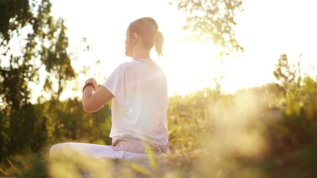 Side view of meditating woman putting hands in namaste mudra posture sitting in lotus pose background of beautiful landscape. Unrecognizable female practicing yoga alone evening sun on nature.