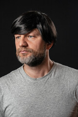 Portrait of attractive man in his 40s with grey and black beard and long black hairs. Black background. Male dressed in grey t-shirt. Looking away from the camera.
