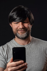 Portrait of a man with grey and black beard and long black hairs. Black background. Male dressed in grey t-shirt, smiling expression on the face, looking at his smart phone.
