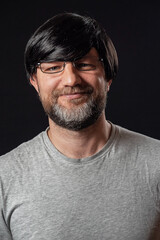 Portrait of a man with grey and black beard and long black hairs. Black background. Male dressed in grey t-shirt, wearing glasses, happy smile expression on his face, looking at the camera