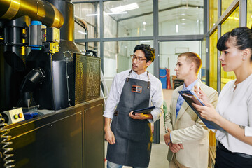 Modern coffee roastery owner in apron showing equipment to inspectors