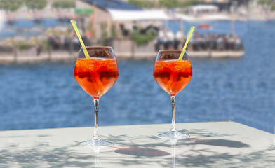 Close up of two glasses of chilled cocktail drinks with straws and  ice cubes on a table near the beach. Blue water and people at a beach restaurant in blurry background.