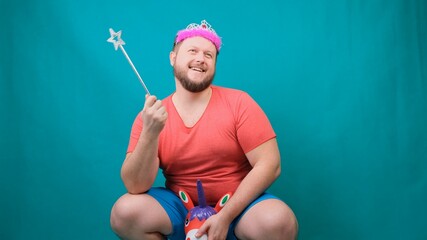 A young bearded freaky man in a pink T-shirt with a diadem on his head riding a unicorn with a magic wand in his hand. A funny wizard joke to make and fulfill a wish.