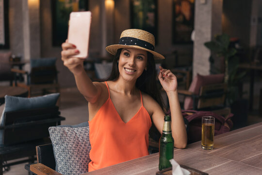 Portrait of young brunette woman taking a selfie photo from cell phone in pub bar. 20s Hispanic girl in summer hat and clothes using mobile phone to video call people in indoors.