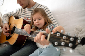 A father teaches a beautiful little daughter to play an acoustic guitar. The child's face looks cute and Funny when she touches the strings.