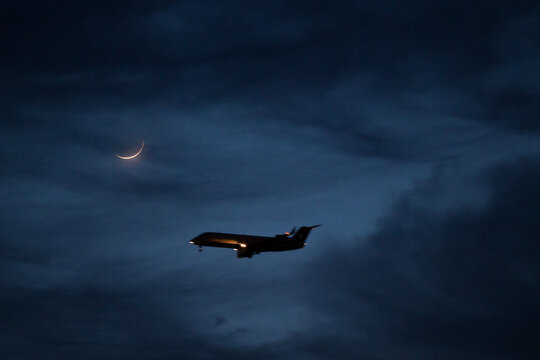 A passenger plane flies against the night sky, lit by the moon