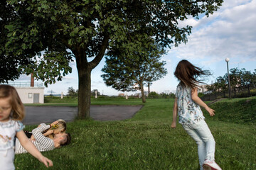 Obraz premium Young parents embrace lying in a green field. Sisters in casual clothes run and play while their parents kiss each other lieing on the grass in a warm summer day.