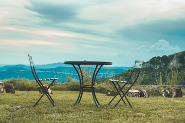 Two chairs facing each other and a table in the middle of nature. Behind it you can see the mountains and the clouds. They are high in a valley.