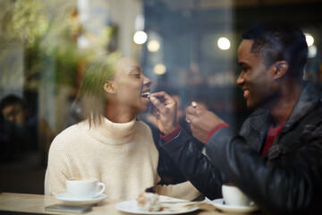 Beautiful couple having lunch, view through cafe window, dark skinned handsome man feeding his woman with dessert cake at their dating