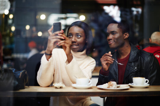 Two dark skinned stylish friends having breakfast in cafe, attractive black woman taking the picture with digital camera on smart phone sitting in beautiful coffee shop, view through cafe window