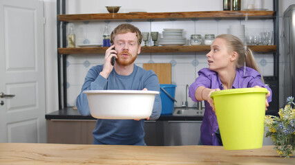 Young couple with pots collecting water falling from ceiling and calling plumber
