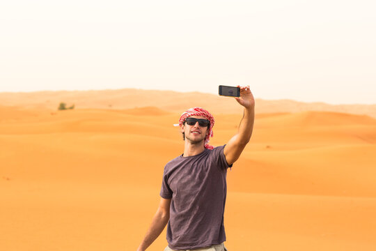 Young male tourist taking a selfie with mobile phone camera in arab desert sand dunes during a safari tour. Dubai tourism and travel adventure.