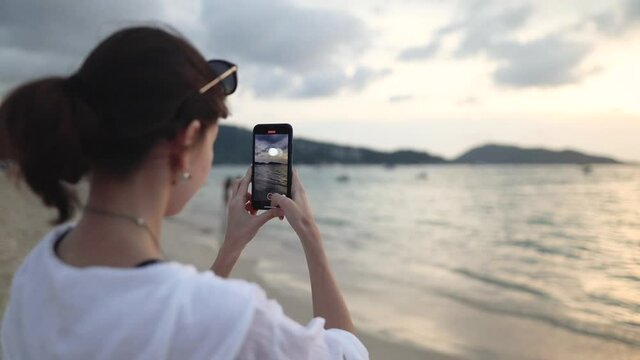 Girl using a smartphone takes a picture of the sunset on Patong Beach, Phuket. In the background is out of focus sea, people passing by. Thailand.