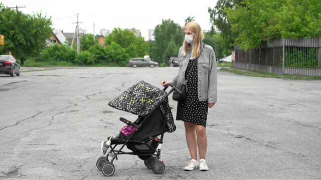 Young mother in a medical mask stands with a stroller in an empty parking lot. Mom is waiting on the street. Concept: covid19, virus, medicine