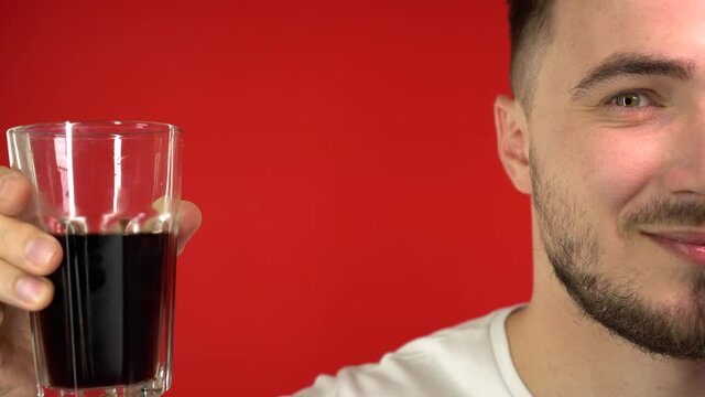 Half the face of a funny man and black soda in a transparent glass glass, the man drinks soda and smiles. Studio videography, on a red background, isolate, 4k.