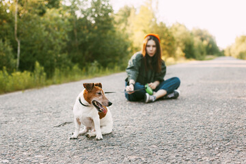 The dog is sitting in front of the girl on the road. A young girl with her pet resting while walking in nature. Blurred background, space for text