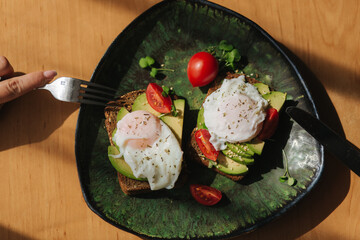 Delicious breakfast at home. Sandwich with fresh sliced avocado above rye toasted bread with cherry tomatoes and poached egg on green plate. Top view