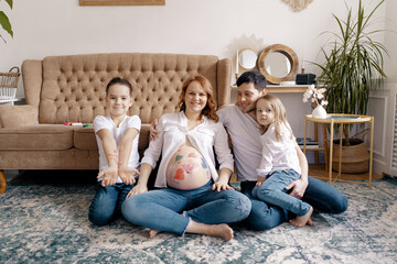 Happy family portrait at home. Parents, pregnant mother and father with lovely two kids sit on the carpet near the sofa. Happy, friendly family. Concept of people and family