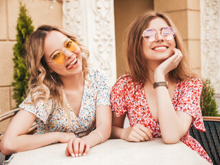 Two young beautiful smiling hipster girls in trendy summer sundress.Carefree women chatting in veranda cafe on the street background in sunglasses.Positive models having fun and communicating