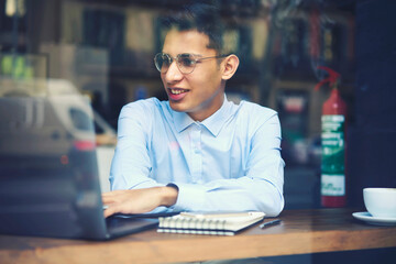 Positive young male student in stylish eyeglasses watching funny video on laptop computer connected to internet.Smiling man reading news in websites and browsing information sitting in cafeteria.