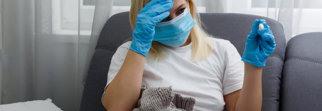 Fever and coronavirus symptoms, woman in medical mask measures body temperature. Sick girl looks at digital thermometer in her hands, concept of cold and flu