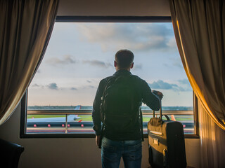 A young man watches the airplane take off from the window of his hotel room.