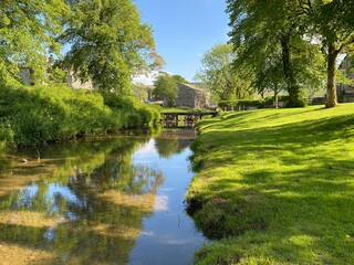 Fototapeta na wymiar Linton Beck, with a green grass sloping bank, old trees and a footbridge, over reflective water in, Linton, Skipton UK