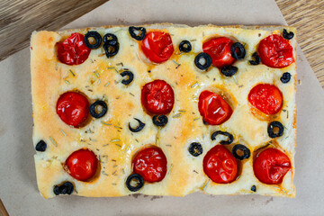 Focaccia with black olives, cherry tomatoes, rosemary and olive oil on a rustic wooden table. 