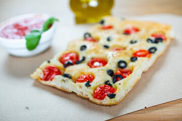 Focaccia with black olives, cherry tomatoes, rosemary and olive oil on a rustic wooden table. 