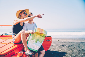 Young hipster girl pointing sitting on rental car explore tropical island with boyfriend on vacations, romantic couple in love discussing direction for sightseeing route during journey by automobile