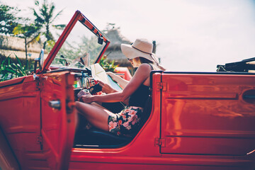 Male and female hipsters sitting in red vintage cabriolet reading map searching direction for...