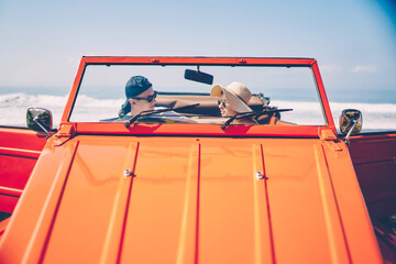 Young hipster couple sitting in rental cabriolet communicating during rest near ocean coast during...