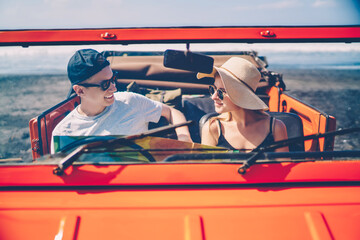 Hipster couple sitting in red rental car resting on ocean beach during roadtrip on summer vacations,smiling female in hat enjoying recreation with boyfriend exploring tropical island on automobile