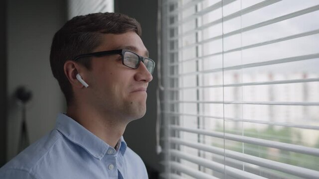 young man with dark hair and glasses in blue shirt stands near window. has phone call uses wireless earphones, nods assent, smiles, feels contented. moves apart jalousies. close up view. side shot
