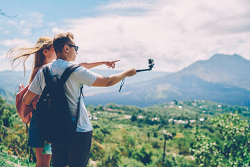 Hipster couple traveling together on summer weekends using monopod for taking selfie,male tourist looking away on beautiful natural environment enjoying recreation trip raking image with girlfriend