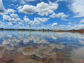 View of salf field under bright blue sky, white fluffy clouds casting reflections over surface of sea water. Salt farming in Thailand.