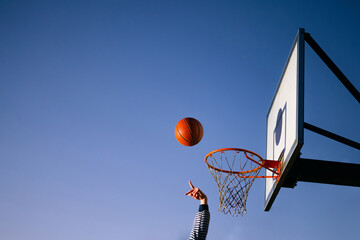 Street basketball ball player throwing ball into the hoop. Close up of hand, orange ball above the hoop net with blue sky in the background. Concept of success, scoring points and winning. Copy space © CrispyMedia