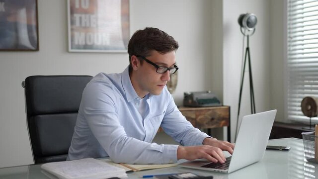 young man in glasses and blue shirt sits at table with laptop in office. is attentive, serious. works with enthusiasm and interest. looks confident and contented. makes stops to think and goes on type