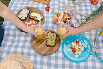 Photo of two hand hold orange lemonade in front of vegan picnic outdoors. Healthy food concept