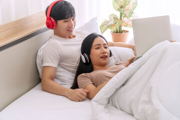 Obraz na płótnie Canvas Cheerful young couple wear wireless headphones enjoy listening music with laptop together on bed in morning. Woman lying on body her boyfriend relax with notebook while looking something at apartment.