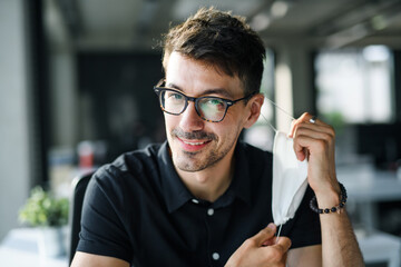 Portrait of young man back at work in office after lockdown, taking off face mask.