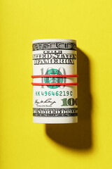 A roll of hundred-dollar American bills is tied with a red elastic band on a yellow background.