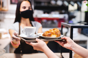 Young woman with face mask working indoors in coffee shop, serving customer.