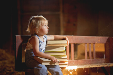 Little toddler boy, sitting on old vintage bench, holding books in attic