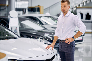 portrait of happy caucasian client man buying new car in car store. luxurious white car is on exhibition in dealership