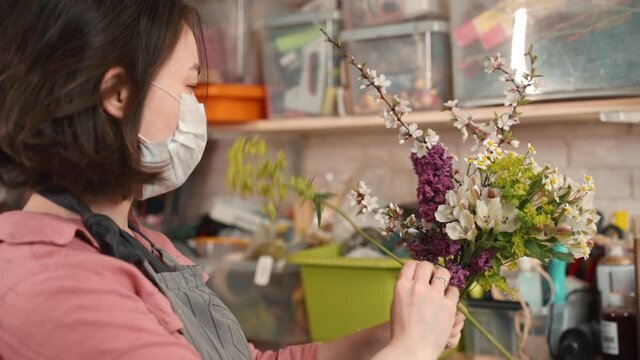 female florist preparing bouquet and wear mask to prevent flu virus. woman working during pandemic in flower shop. making composing floral decoration. concept virus, prevention, distancing.