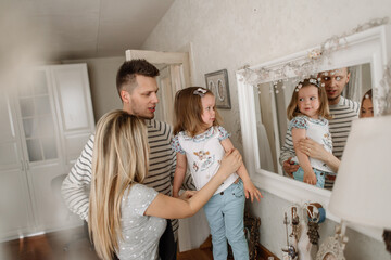 Two parents with their little beautiful daughter look in a vintage mirror. The child explores her reflection. The family is isolated at home and looks protected and calm.