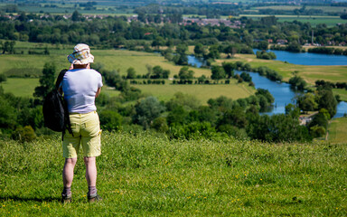 Fototapeta premium An older woman photographing a landscape in Wittenham Clumps, carrying backpack, enjoying the weather, lady wearing blue t-shirt, creamy shorts and hat, countryside stroll with river view
