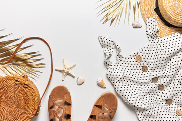 Summer women's white dress in black peas rattan woven bag brown sandals straw hat golden palm leaf shells starfish on light background. Flat lay top view. Women's beach fashion, travel vacation