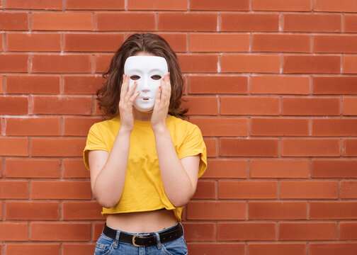 Young Woman In Yellow Shirt Putting On A Mask Against Brick Wall